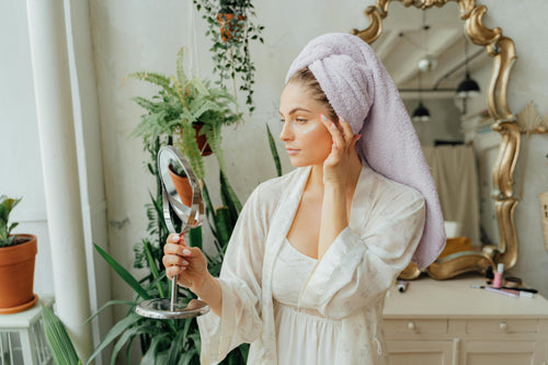 Woman in robe and towel applying face cream with mirror