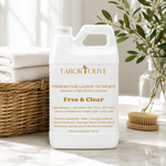 Load image into Gallery viewer, White bottle of Tabor and Olive Premium Free and Clear Liquid Laundry Detergent on a marble platform next to a basket of white towels and decor.
