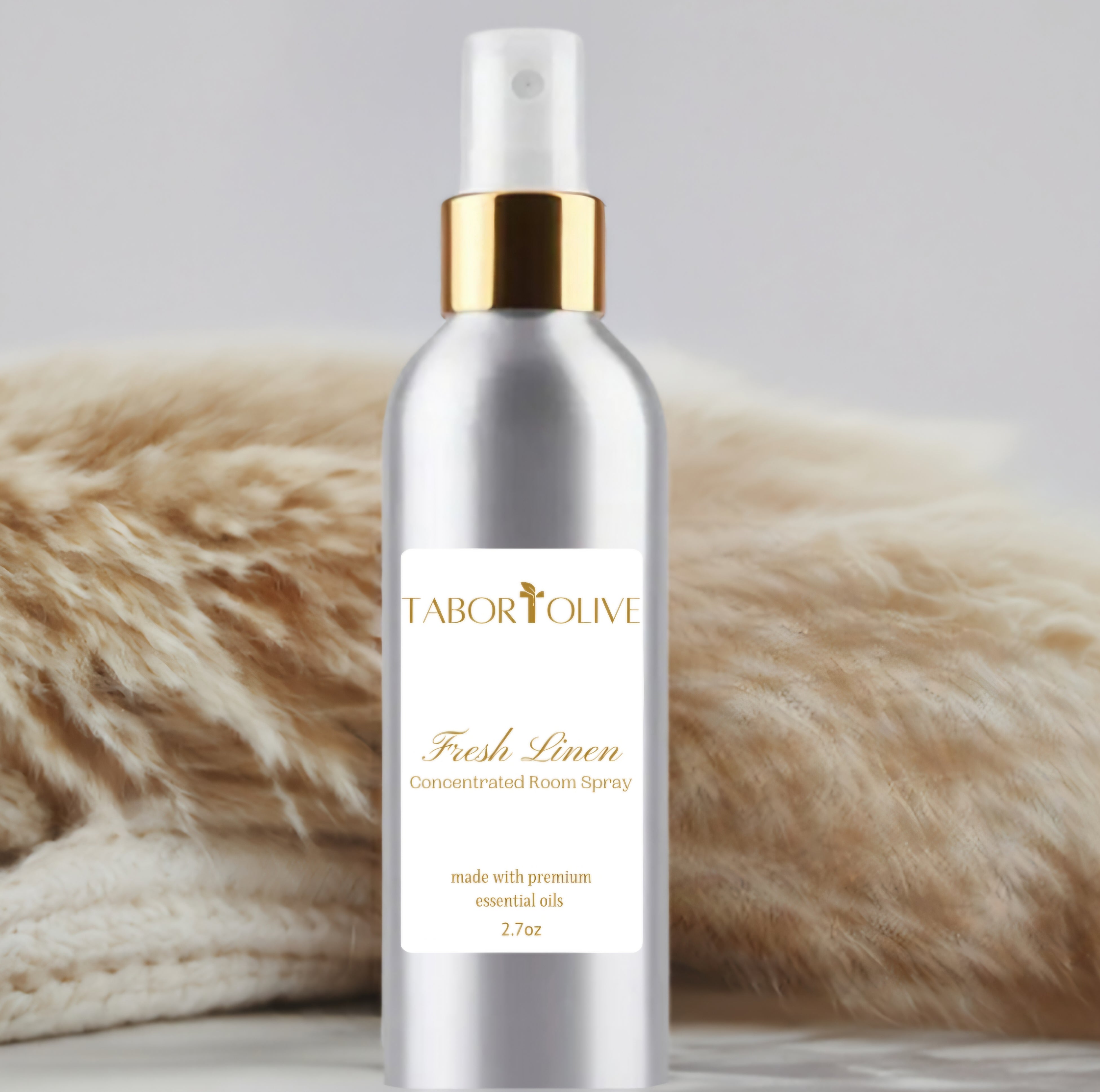 Aluminum spray bottle with gold cap sitting on marble platform next to pampas grass and a blanket that says,  "concentrated room spray. Made with premium essential oils." 