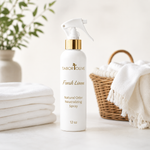 Load image into Gallery viewer, A 12 oz white spray bottle of Tabor Olive Fresh Linen Natural Odor Neutralizing Spray with a gold collar and white trigger sprayer, set on a white surface with folded white towels, a wicker basket, and a potted plant in the background.
