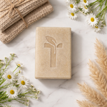 Load image into Gallery viewer, Beige soap bar with a leaf design on a marble surface with flowers and towels.
